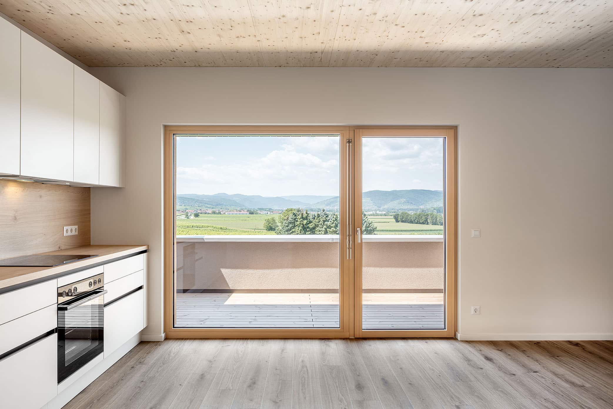 Ansicht Küche in einem Massivholzwohnbau mit großem Fenster und Terrassentür, Parkettboden, Sichtholzdecke